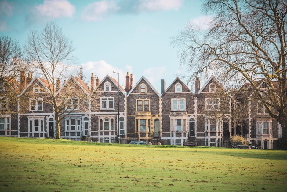 A row of three-storey Victorian-style terraced houses with dark grey brick facades, white window frames, and pitched roofs, situated along a residential street. In the foreground, there is a grassy pa