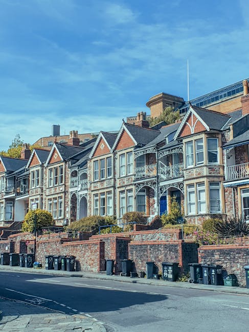 A row of three-storey Victorian-style terraced houses with dark grey brick facades, white window frames, and pitched roofs, situated along a residential street. In the foreground, there is a grassy pa