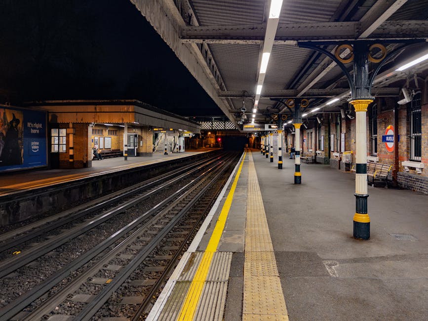 An empty London Underground station platform at night, with the platform surface illuminated by overhead lighting. The platform has tactile paving along the edge, yellow safety lines, and is supported by ornate, black and yellow decorative columns. On the right side, there are brick walls with windows, some benches, and signage indicating the station's name and safety instructions. The tracks run down the center, leading into the dark tunnel at the far end. This scene depicts a quiet moment during night-time, with no passengers visible, highlighting the infrastructure typical of London transport hubs, suitable for a context involving house removals or furniture transport services such as those offered by Finsbury Park Man and Van.