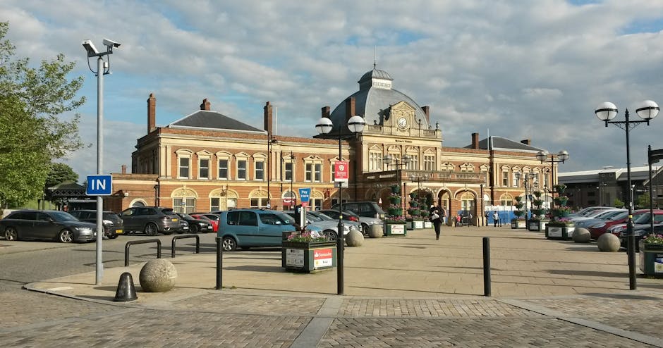 A spacious public square in front of a historic station building featuring Victorian-style architecture with ornate detailing, arched windows, and a central clock tower topped with a small dome. The area is paved with concrete, with black metal bollards and wheel stops positioned along the perimeter. Several parked cars, including sedans and hatchbacks, are arranged in neat rows across the parking area adjacent to the station. Flower planters with colorful blooms are placed near the entrance and along the edges of the square, which is illuminated by tall street lamps with double-globe fixtures. A few pedestrians are visible walking across the open space, approaching the station entrance. The scene is captured during daylight hours, with a partly cloudy sky overhead, and the overall environment appears organized and well-maintained, suitable for a home relocation or furniture transport process involving Finsbury Park Man and Van's removals services.