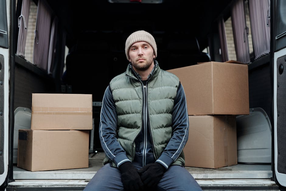 A man dressed in a blue beanie, black track suit with red sleeves, and a dark jacket is placing two cardboard boxes onto the ground next to a silver panel van parked in a designated loading area outsi