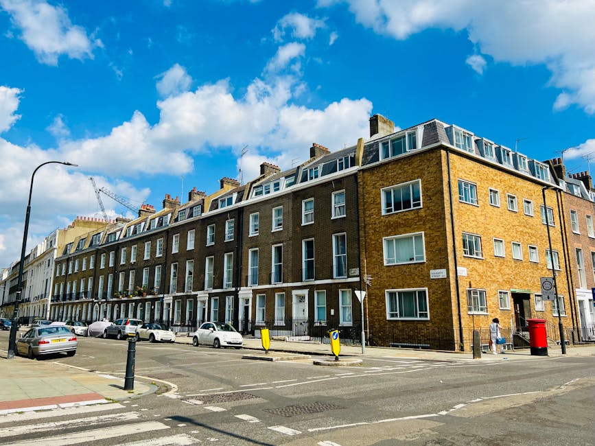 A street scene featuring a large, multi-storey brick residential building with several rows of white-framed double-glazed windows, some with window boxes and curtains. The building's ground floor includes doorways with steps, and a black iron fence runs along the sidewalk in front. Parked cars of various sizes and colors line the curb on both sides of the street, with a pedestrian crossing visible in the foreground. On the sidewalk, a person with a backpack approaches a red post box beside a lamppost, while a construction crane is visible in the distance under a bright blue sky with scattered white clouds. The atmosphere suggests a typical urban environment suitable for house or flat relocations, with potential involvement of professional removals services like Finsbury Park Man and Van during moving logistics.