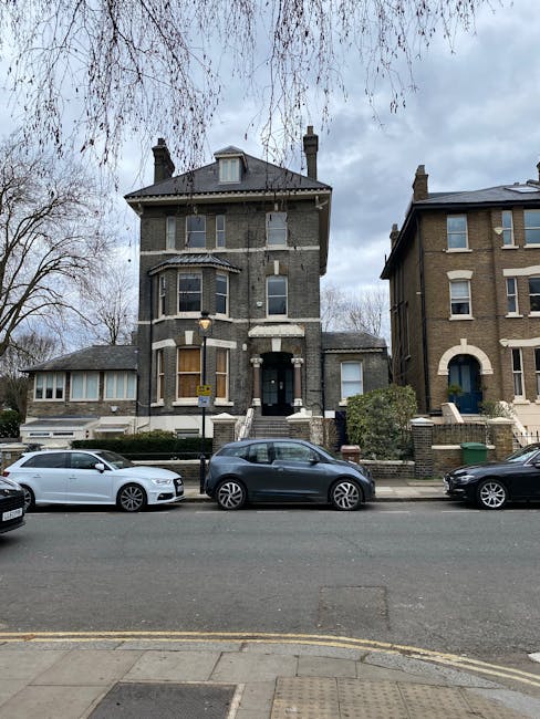 A three-storey Victorian-style house on Stroud Green Road N4, Finsbury Park, facing the street. The building features a dark brick facade with decorative stone window surrounds and a central entrance accessed by a short flight of steps. To the left, there is a smaller, single-storey extension with a large window. Three cars are parked along the pavement directly in front of the house, including a white, a grey, and a black vehicle. The street is lined with leafless trees and a cloudy sky overhead. The overall scene captures the residential environment during daytime, focusing on the housing style and the street's parking area, consistent with house removal and relocation services offered by Finsbury Park Man and Van.