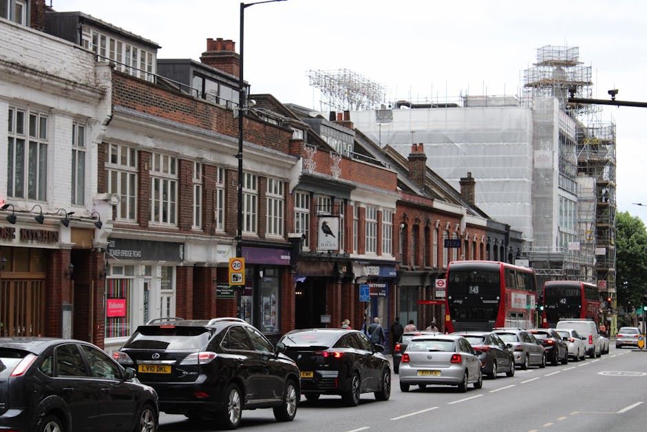 The image depicts a busy street scene on Seven Sisters Road N4 in Finsbury Park, showing a line of parked cars and two red double-decker buses moving along the road. The storefronts and commercial units occupy the ground level of a row of traditional brick buildings, some with white window frames and decorative facade elements. A construction site with scaffolding and protective sheeting is visible at the far end of the street, indicating ongoing building work. Near the curb, a team of movers from Finsbury Park Man and Van is engaged in a home relocation process, loading or unloading furniture and packed cardboard boxes onto a van, which is partially visible on the street. The movers use equipment such as trolleys and moving blankets to secure and transport household items, reflecting professional packing and furniture transport activities typical during house removals in urban areas. The environment is well-lit with natural daylight, and pedestrians are seen walking along the pavement, contributing to the busy urban atmosphere of the street.