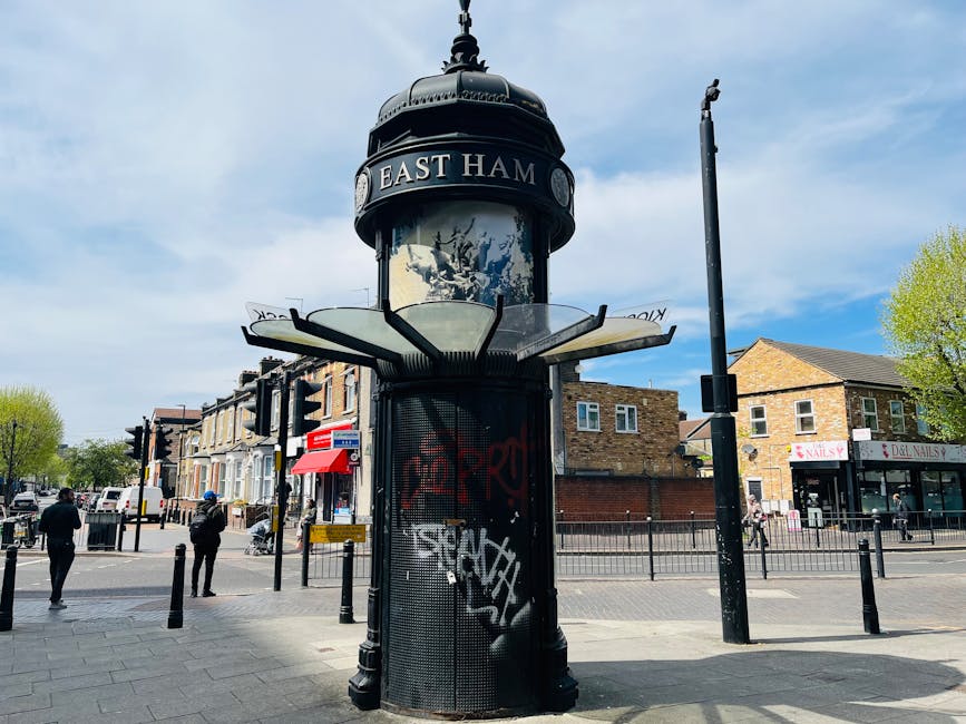 A tall, black, historical-looking public telephone kiosk with a domed, decorative top featuring a circular sign that reads 'EAST HAM.' The kiosk has a glass window section below the sign, photoed on a city street corner during daytime with a partly cloudy sky. Surrounding the kiosk are pedestrians walking on the pavement, some of whom are wearing dark clothing. In the background, there are low-rise residential and commercial buildings made of brick and stone, including a nail salon, shops with red and white signage, and a bus stop with a red canopy. The street features black metal bollards along the pavement edge, traffic lights, and a street lamp. The scene is well-lit with natural sunlight, casting shadows on the ground and highlighting the urban environment consistent with a typical cityscape suitable for house removals or furniture transport activities carried out by professional services such as Finsbury Park Man and Van.